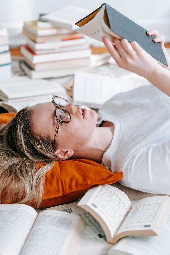 Side view of focused female in eyeglasses reading story in book while lying on floor amidst heap of literature in room
